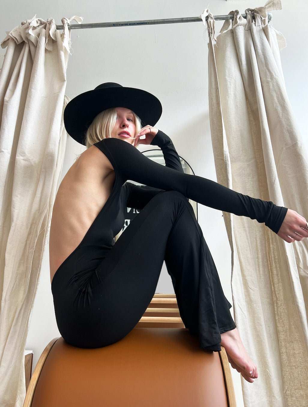 The Bond Bodysuit black fitted one-piece with open back and flare legs worn with black hat