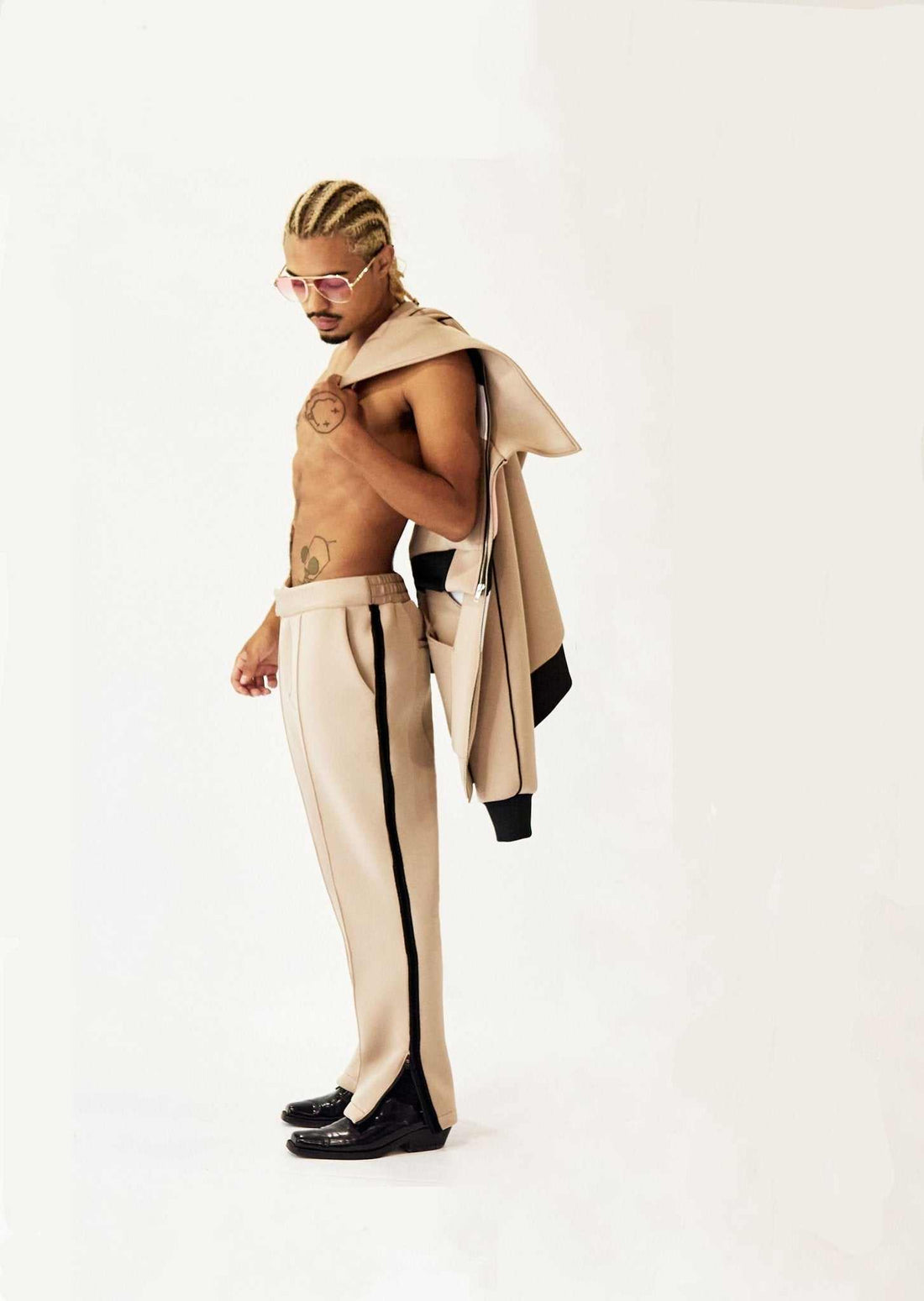 Man wearing beige Tuxedo Track Pant with velvet side tape and ankle zippers, holding matching jacket over shoulder.