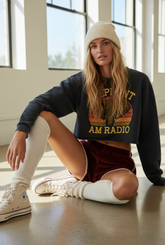 Woman wearing black ELEPHANT CROP SWEATSHIRT with vintage logo, knit beanie, and casual shorts seated indoors
